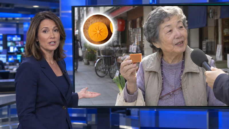 A news anchor in a studio in front of a screen showing a street interview with an elderly Japanese woman holding a bar of persimmon soap