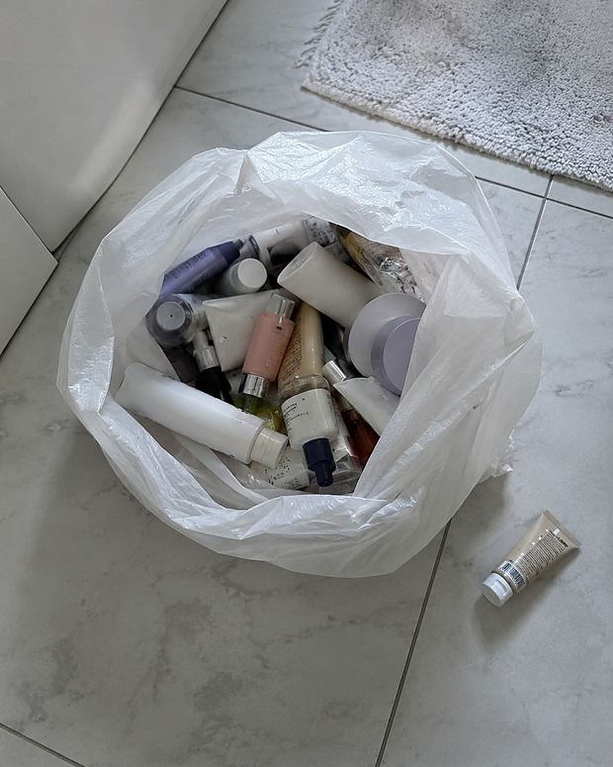 A trash bag on a bathroom floor full of old cream tubes and powder bottles